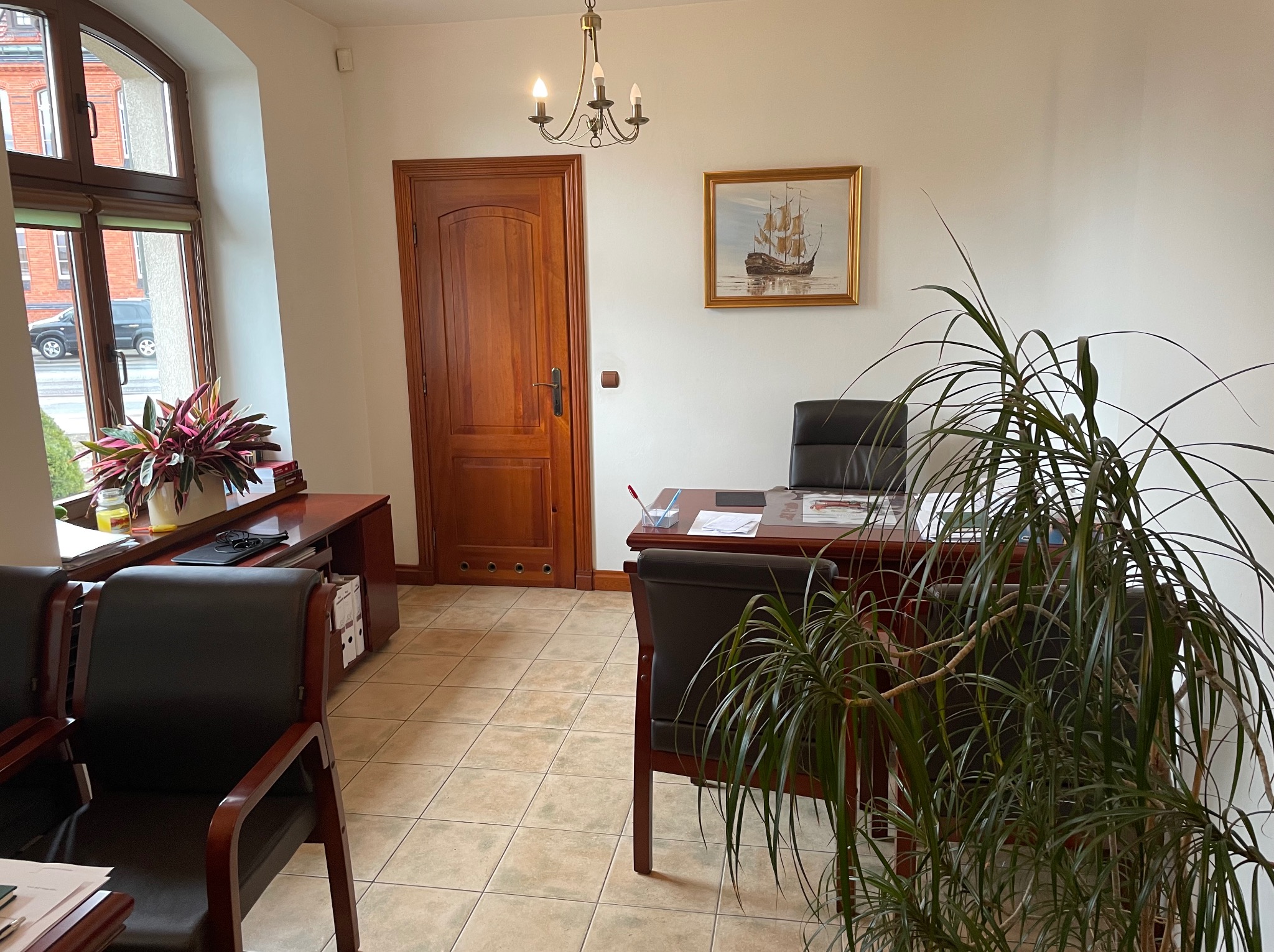 Eleganckie biuro: drewniane meble, obraz z żaglowcem, roślina doniczkowa. Stonowana kolorystyka, klasyczny wystrój. Widok na ulicę zza okna.