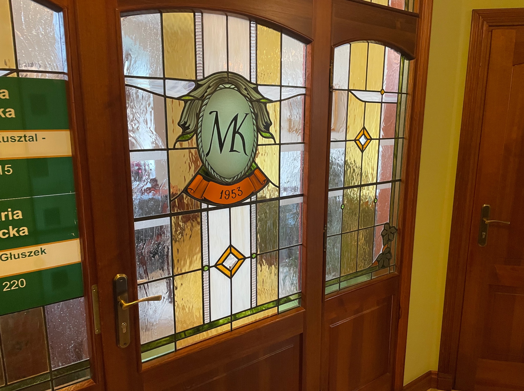 Stylowe, drewniane drzwi z witrażem z inicjałami 'MK' i datą 1955 w zielono-pomarańczowej kolorystyce, ozdobione geometrycznymi wzorami i motywami roślinnymi.