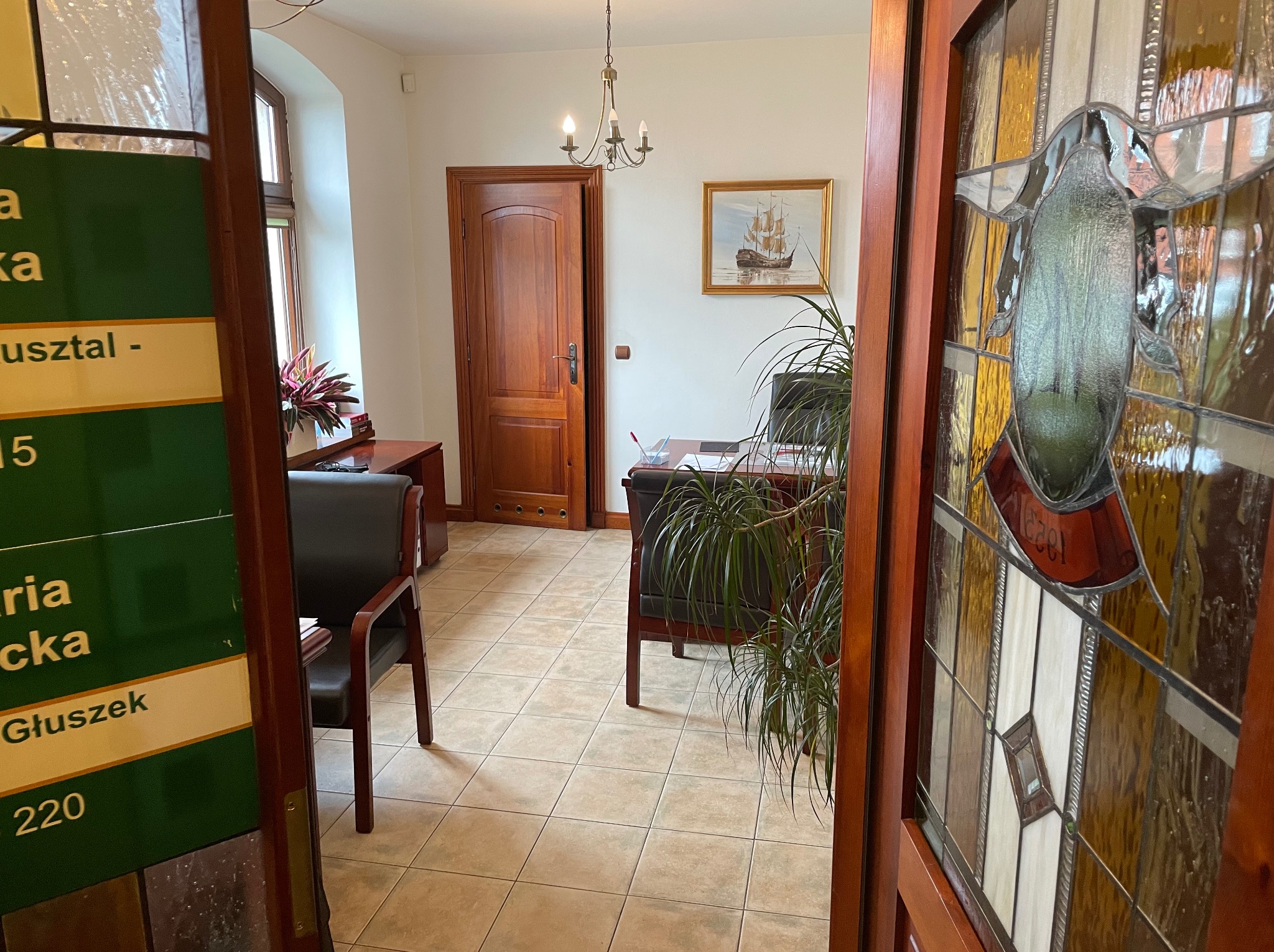 Kancelaria prawna z drewnianymi drzwiami i witrażem w tle. Widoczny fragment biura, krzesła i biurko. Na ścianie obraz z motywem żaglowca.