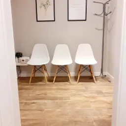 Poczekalnia z trzema białymi krzesłami Eames, w tle grafiki w czarnych ramkach i wieszak na ubrania.