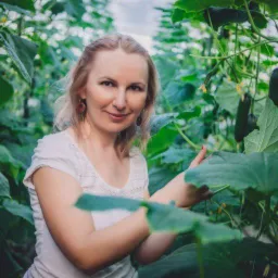 Kobieta w jasnej koszulce w szklarni z og&oacute;rkami, trzymająca liść, z widocznymi owocami i kwiatami.