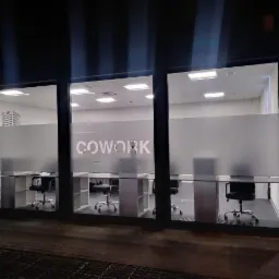 Elewacja budynku z przeszklonymi oknami, za którymi widoczne stanowiska pracy w przestrzeni typu cowork z biurkami, krzesłami i logo 'COWORK' na matowej folii okiennej.
