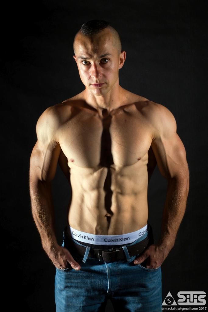 Mężczyzna z wyraźnie zarysowanymi mięśniami brzucha, ramion i klatki piersiowej, pozujący w bieliźnie marki Calvin Klein i dżinsach na ciemnym tle.