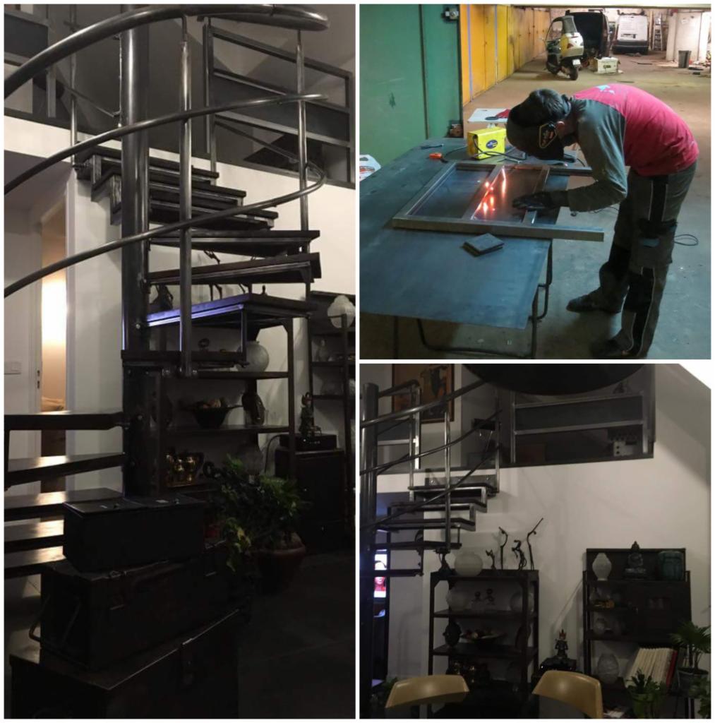 Metalowa spiralna klatka schodowa w pomieszczeniu mieszkalnym, obok regały z dekoracjami; w tle spawanie metalowej ramy okiennej przez osobę w masce ochronnej.