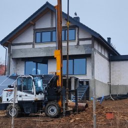 Dewax Sp.z o.o. - Żółta wiertnica hydrauliczna na tle domu w trakcie budowy, obok panele słoneczne i częściowo widoczny mężczyzna w kurtce