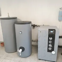 Instalacja systemu grzewczego w Dobrzycy: dwa szare zasobniki ciepłej wody i jednostka sterująca z folią ochronną, widoczne rury i armatura, na ścianie skrzynka elektryczna.