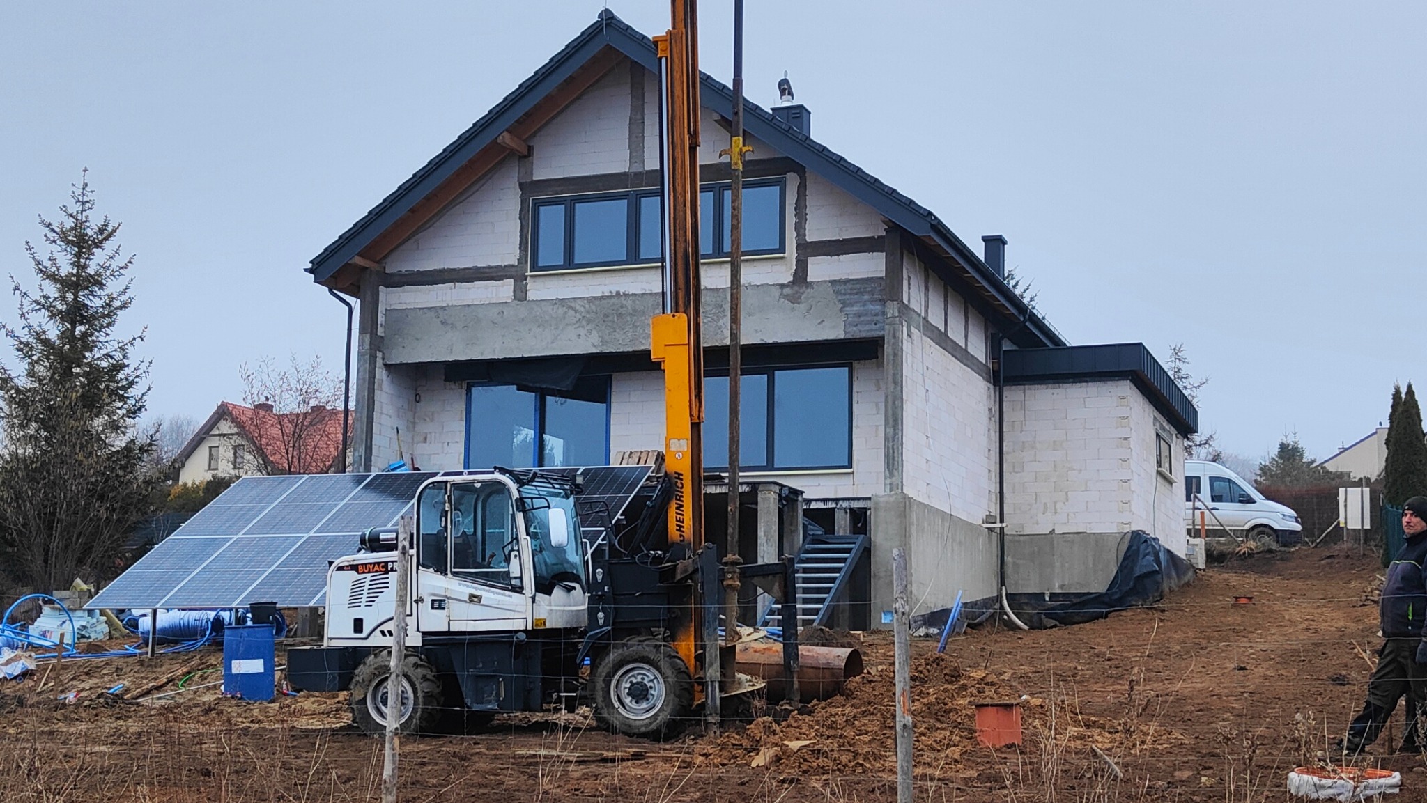Żółta wiertnica hydrauliczna na tle domu w trakcie budowy, obok panele słoneczne i częściowo widoczny mężczyzna w kurtce