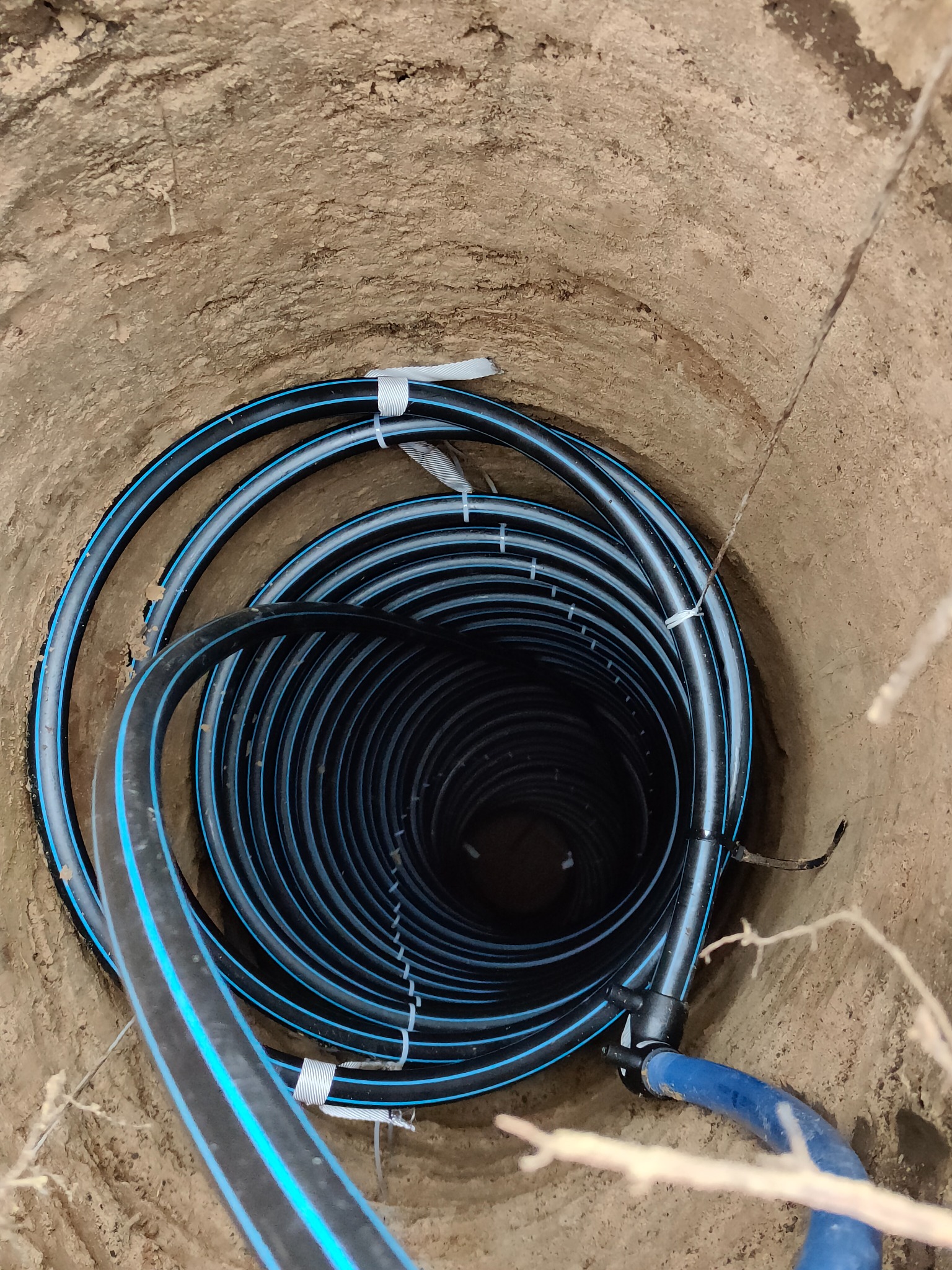 Wykop w ziemi z ułożonym w spiralę czarnym przewodem z niebieskimi paskami, zabezpieczonym białymi opaskami zaciskowymi, widok z góry.