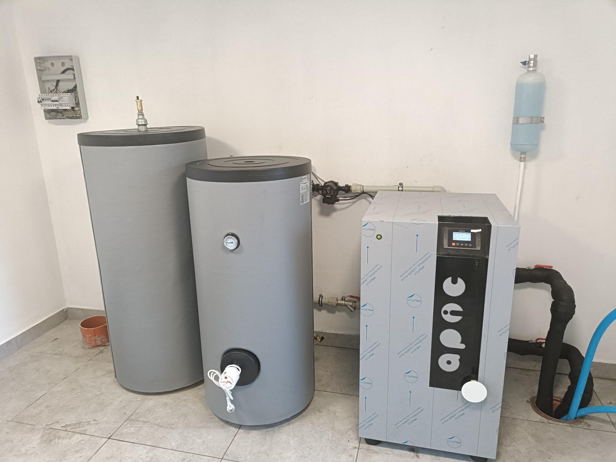 Instalacja systemu grzewczego w Dobrzycy: dwa szare zasobniki ciepłej wody i jednostka sterująca z folią ochronną, widoczne rury i armatura, na ścianie skrzynka elektryczna.