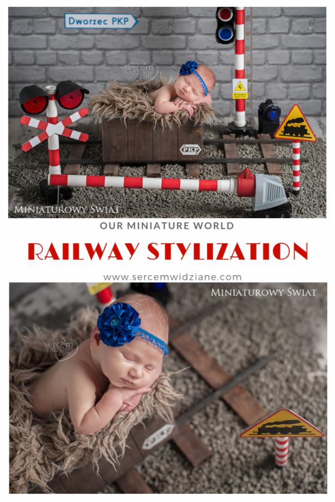 Noworodek śpiący w drewnianej skrzynce imitującej wagon kolejowy, na tle miniaturowej scenografii kolejowej z przejazdem, semaforem i znakiem ostrzegawczym, całość w tonacji szarości i brązu.