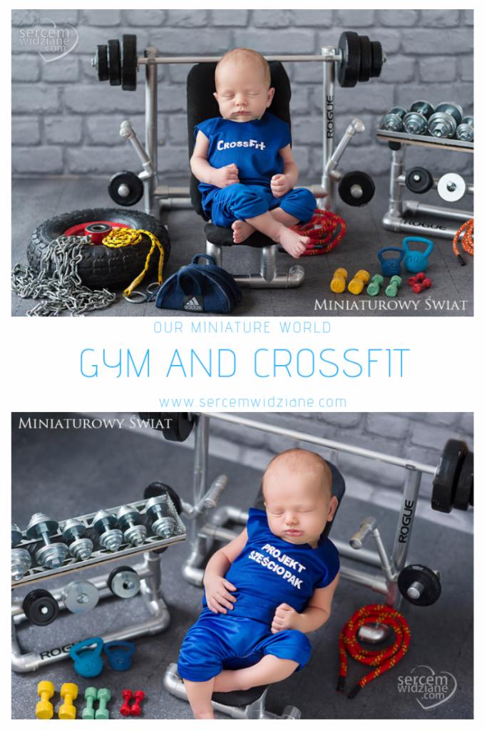 Śpiący noworodek ubrany w niebieski strój sportowy z napisem 'CrossFit' siedzi na miniaturowej ławeczce do ćwiczeń otoczony miniaturowym sprzętem do ćwiczeń, w tle ściana z cegieł.
