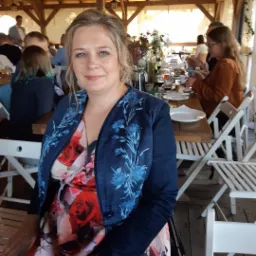 Portret kobiety w kwiecistej sukience i granatowej marynarce z niebieskimi kwiatami na tle stołu z nakryciami i siedzących osób w plenerowej restauracji.