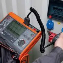 Miernik wielofunkcyjny w pomarańczowej obudowie z czarnym przewodem podłączony do gniazda w skrzynce rozdzielczej podczas wykonywania pomiarów przez elektryka w szarym ubraniu roboczym.