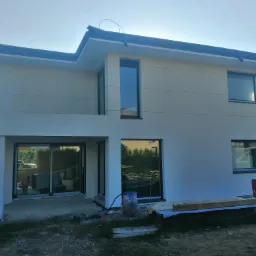 Nowoczesny dom jednorodzinny z jasną elewacją i ciemną stolarką okienną, widoczne elementy rusztowania i materiały budowlane, sugerujące prace wykończeniowe.