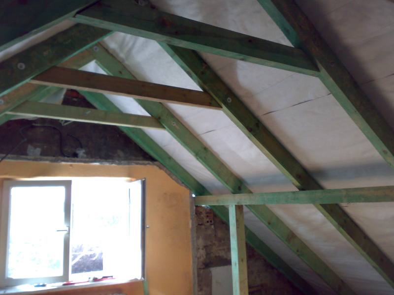 Konstrukcja drewniana dachu z widoczną membraną dachową, surowy stan poddasza z oknem, elementy pomalowane na zielono.