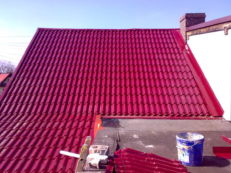 Czerwony dach z blachodachówki, widoczny z góry, z fragmentem muru kominowego i niebieskim wiadrem na dachu sąsiedniego budynku.