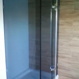 Kabina prysznicowa z ciemnego szkła, z chromowanymi uchwytami, zamontowana w rogu łazienki wyłożonej płytkami imitującymi drewno.