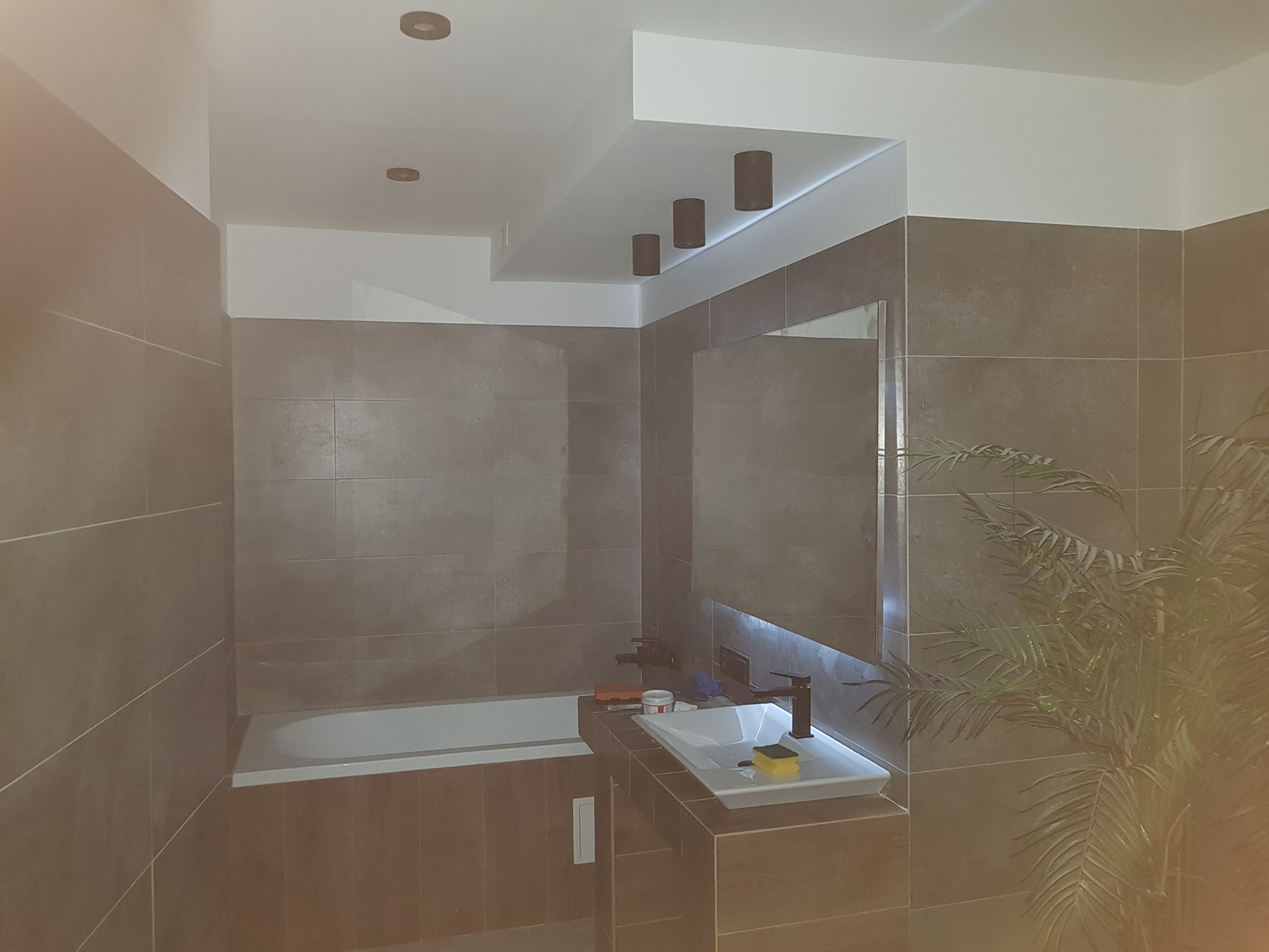 Nowoczesna łazienka z brązowymi płytkami, białym sufitem z wpuszczanymi reflektorami, białą wanną i umywalką z czarną armaturą, oraz rośliną doniczkową.