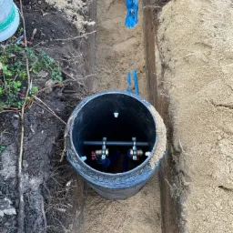 Wykop z czarną studzienką rewizyjną z podłączeniem rur, widoczne zawory kulowe z czerwonymi rączkami, niebieskie rury w ziemi, obok ziemia z korzeniami i ostrzegawcza niebieska folia.