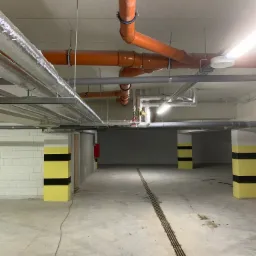 Widok na sufit garażu podziemnego z widocznymi pomarańczowymi rurami instalacji i srebrnymi rurami wentylacyjnymi, poprowadzonymi wzdłuż betonowego sufitu, z kolumnami pomalowanymi w żółto-czarne...