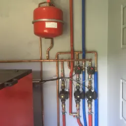 Czerwony zbiornik wyrównawczy z manometrem, miedziane rury instalacji grzewczej, pompy obiegowe oraz izolowane rury w kolorach czerwonym i niebieskim, zamontowane na szarej ścianie.