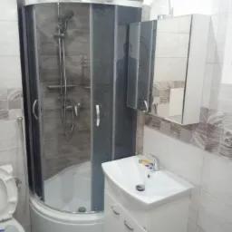 Nowoczesna łazienka z kabiną prysznicową z grafitowymi szybami, białą umywalką z szafką i lustrem z oświetleniem, wykończona płytkami w odcieniach szarości i beżu.