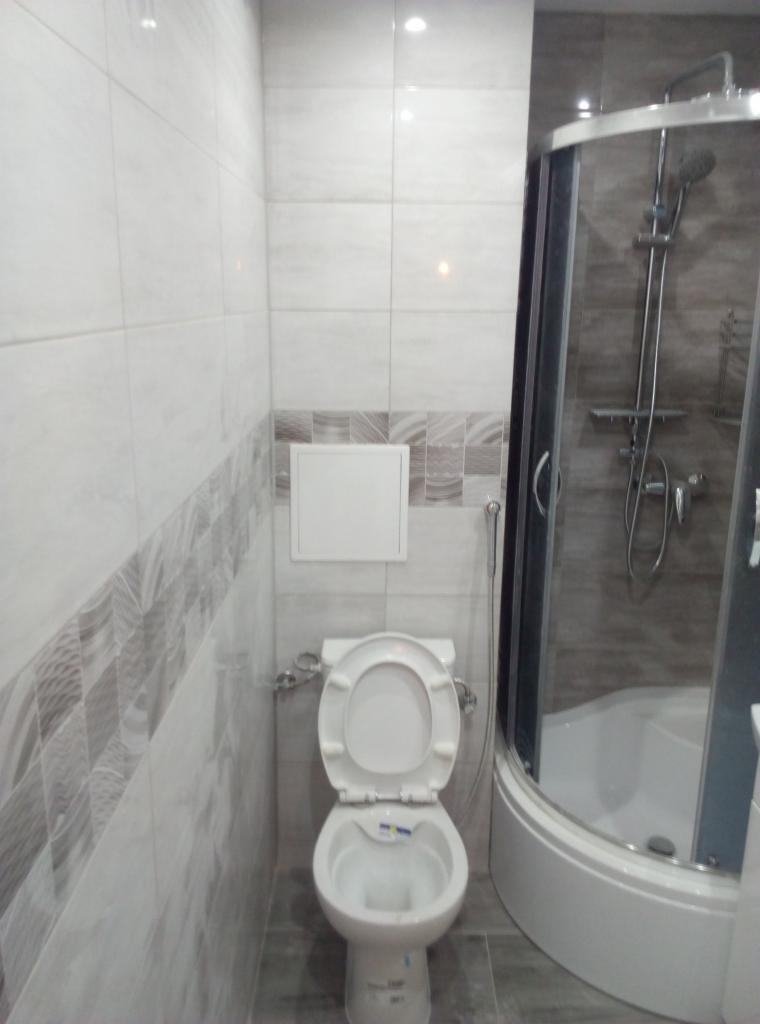 Wykończona łazienka z białą toaletą z otwartą klapą, kabiną prysznicową z ciemnymi ramami i szarymi płytkami z dekoracyjnym pasem.