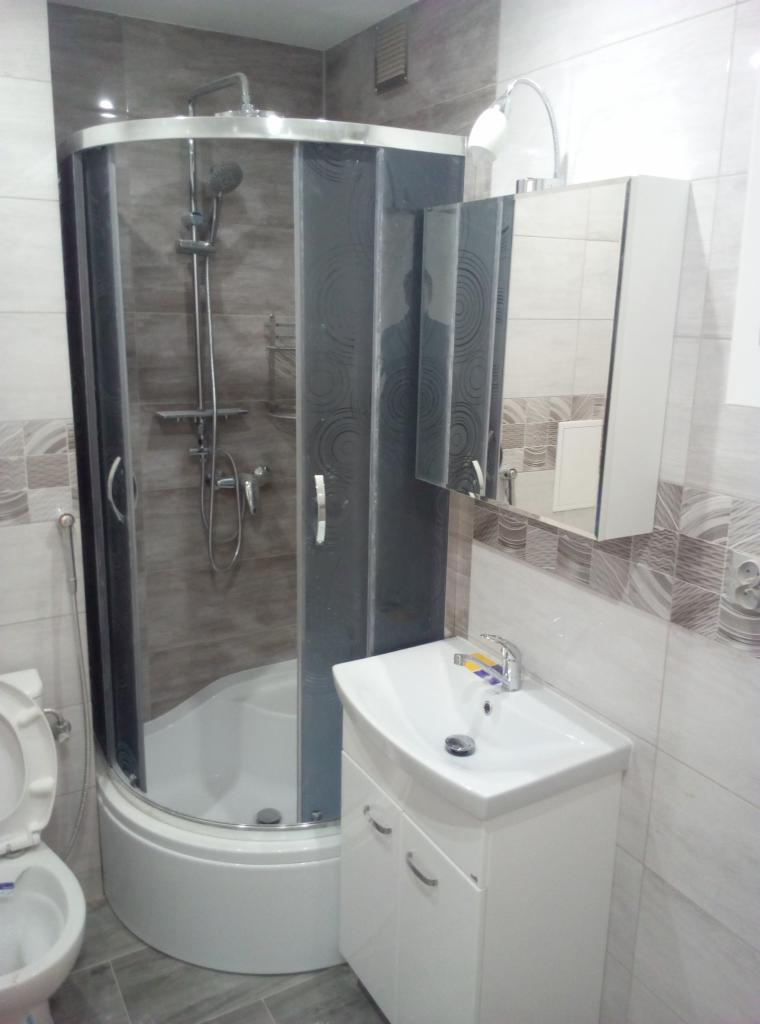 Nowoczesna łazienka z kabiną prysznicową z grafitowymi szybami, białą umywalką z szafką i lustrem z oświetleniem, wykończona płytkami w odcieniach szarości i beżu.