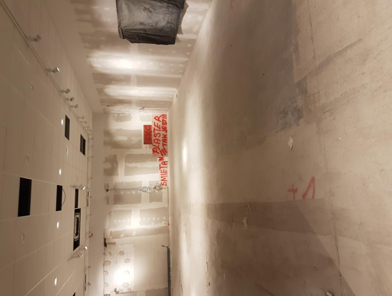 Wnętrze w trakcie remontu: ściany przygotowane pod gładzie, widoczne napisy 'Śmietan Plaster Tak Jest!!!' wykonane czerwoną farbą, sufit z zamontowanymi punktami oświetleniowymi.