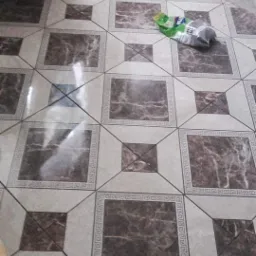 Ułożona podłoga z płytek w odcieniach brązu i beżu z dekoracyjnym wzorem greckim wokół większych kwadratów, widoczny śmieć na podłodze.