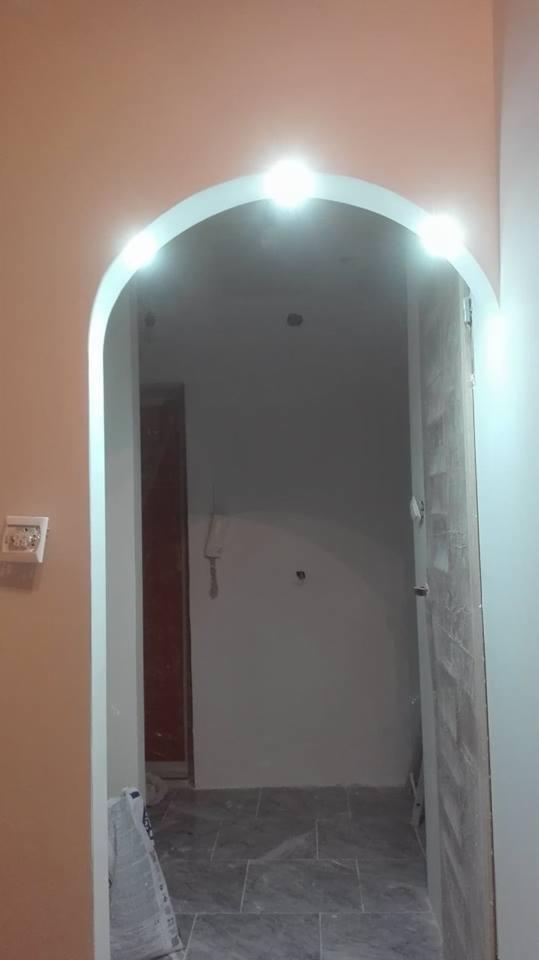 Łukowate przejście z zamontowanym oświetleniem LED, szare płytki podłogowe, ściany w odcieniach beżu i szarości, widoczne ślady po remoncie, częściowo otwarte drzwi.