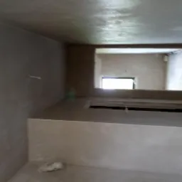 Surowe, świeżo nałożone tynki na ścianach i suficie w pomieszczeniu z widocznym otworem okiennym w oddali, ukazujące nierówności i fakturę powierzchni przed wygładzeniem.