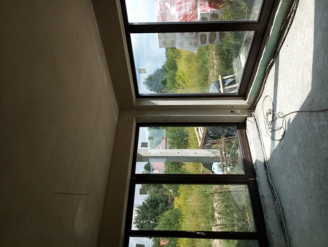 Wnętrze surowego budynku w trakcie prac wykończeniowych: ściany po nałożeniu gładzi, widoczne okna z brązowymi ramami i widok na zewnątrz, kable elektryczne na podłodze.