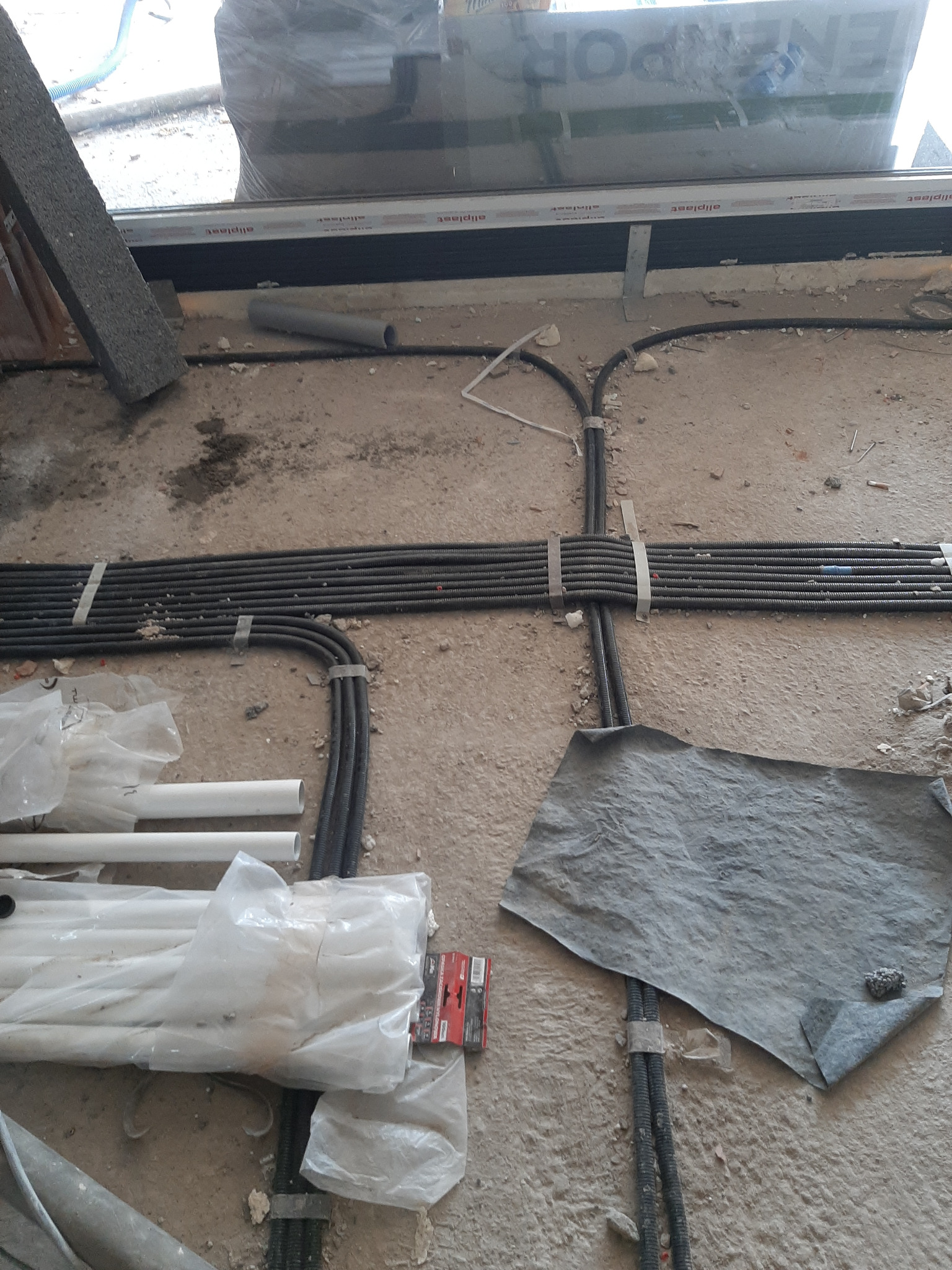 Wiązki przewodów elektrycznych ułożone na surowej podłodze, częściowo przykryte folią ochronną, widoczne także rury PCV w folii i okno w trakcie montażu.