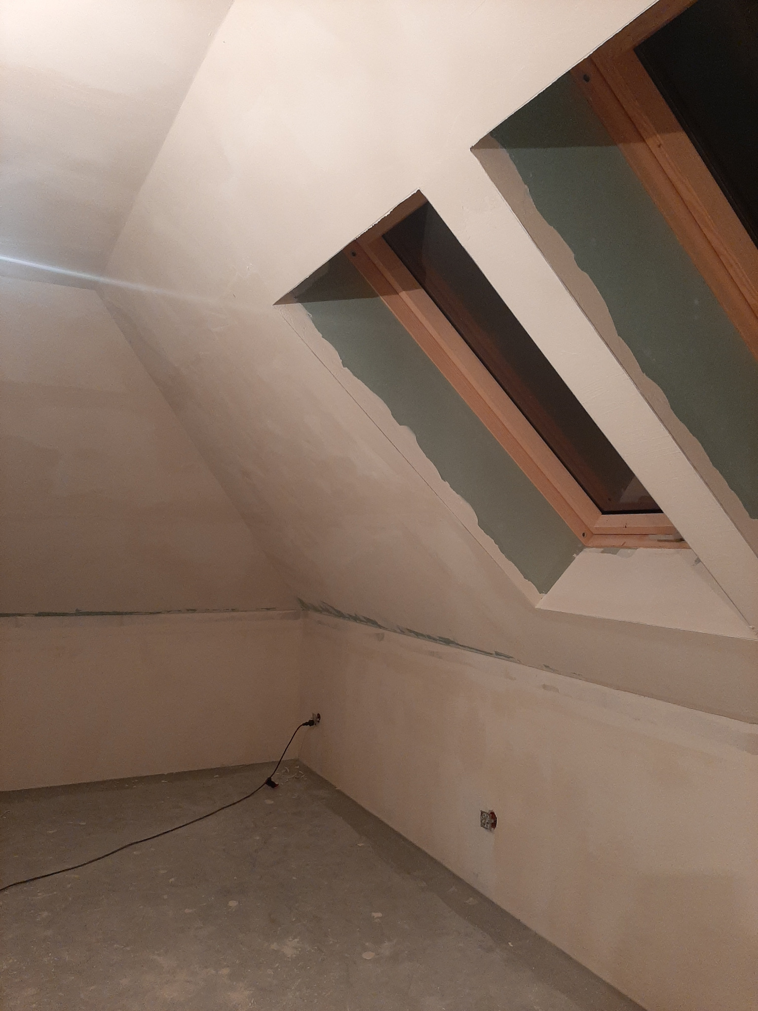 Poddasze w trakcie szpachlowania, widoczne dwa okna dachowe z drewnianą ramą, ściany i sufit pokryte gładzią szpachlową, widoczne gniazdka elektryczne i przedłużacz na podłodze.