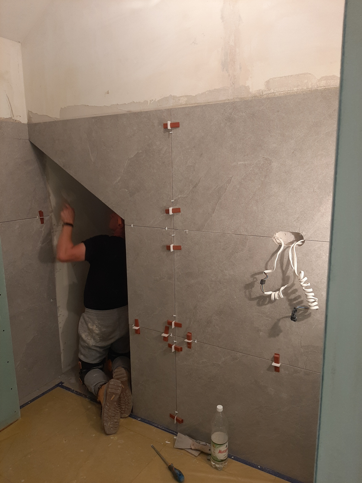 Pracownik na kolanach układa szare płytki w łazience, widoczne krzyżyki dystansowe, otwór na instalację elektryczną i butelka wody mineralnej na podłodze.