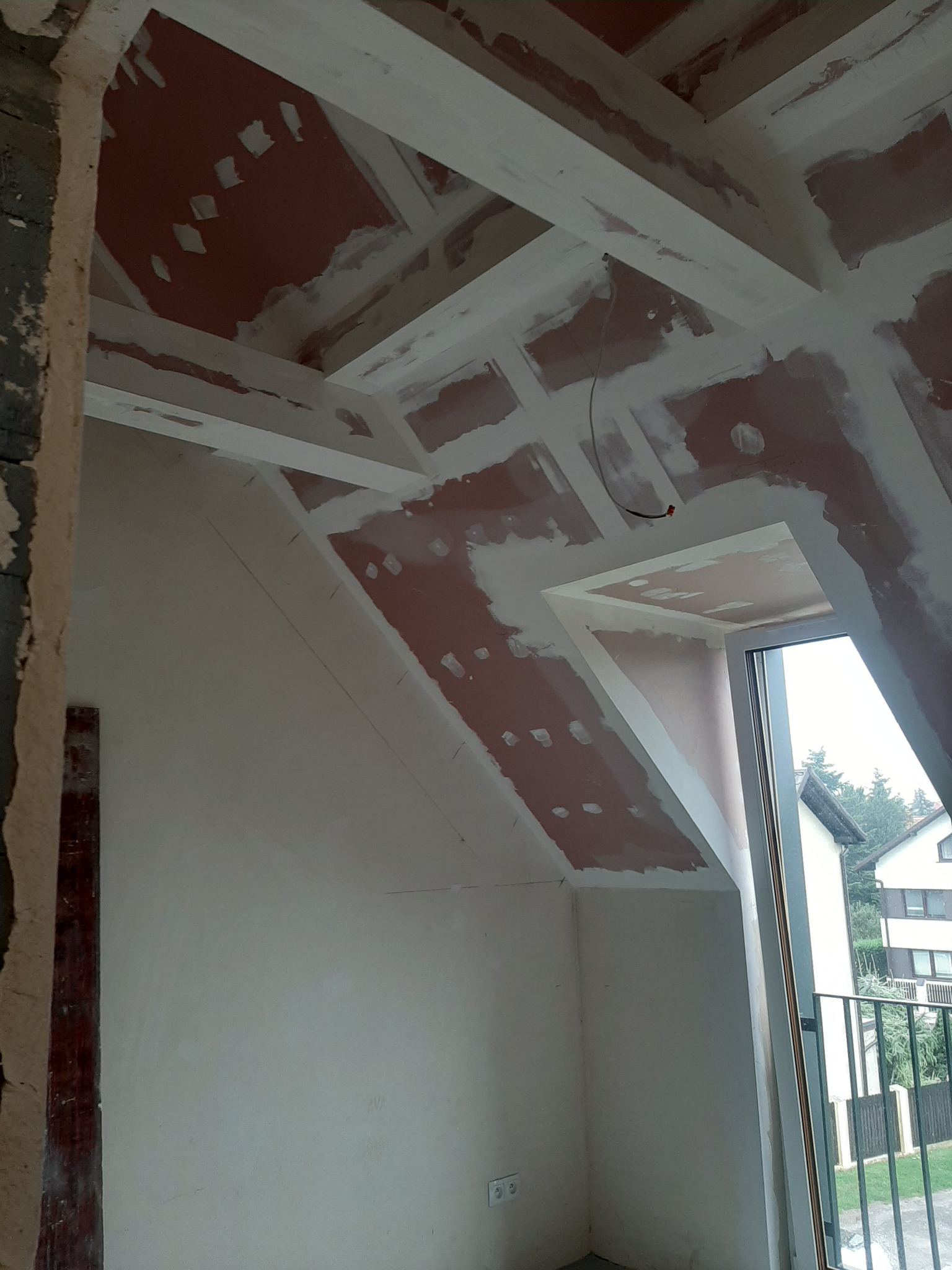 Kraków: Wnętrze poddasza w trakcie remontu, widoczne surowe ściany z nałożoną gładzią szpachlową i drewniane belki stropowe przygotowane do malowania, okno dachowe z widokiem na zieleń.