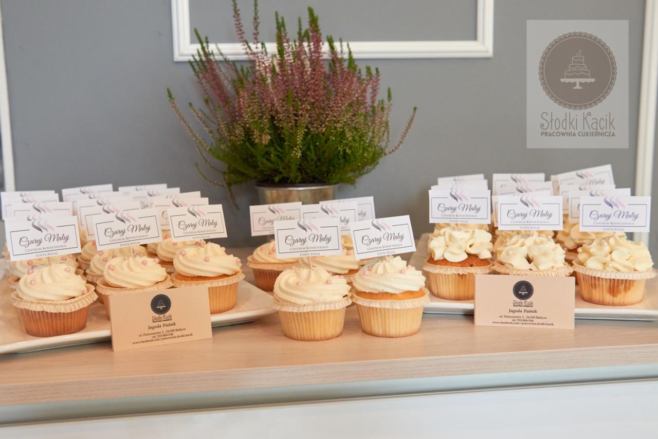 Rząd babeczek z kremem i różowymi posypkami, za nimi wizytówki z nazwą firmy oraz wrzos w metalowej doniczce na jasnym blacie.