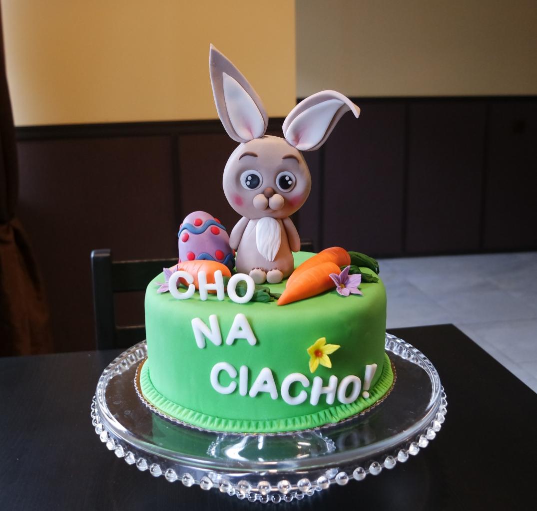 Okrągły tort z zielonym lukrem, dekorowany figurką zajączka, pisanką, marchewkami i napisem 'CHO NA CIACHO!', umieszczony na szklanej paterze.
