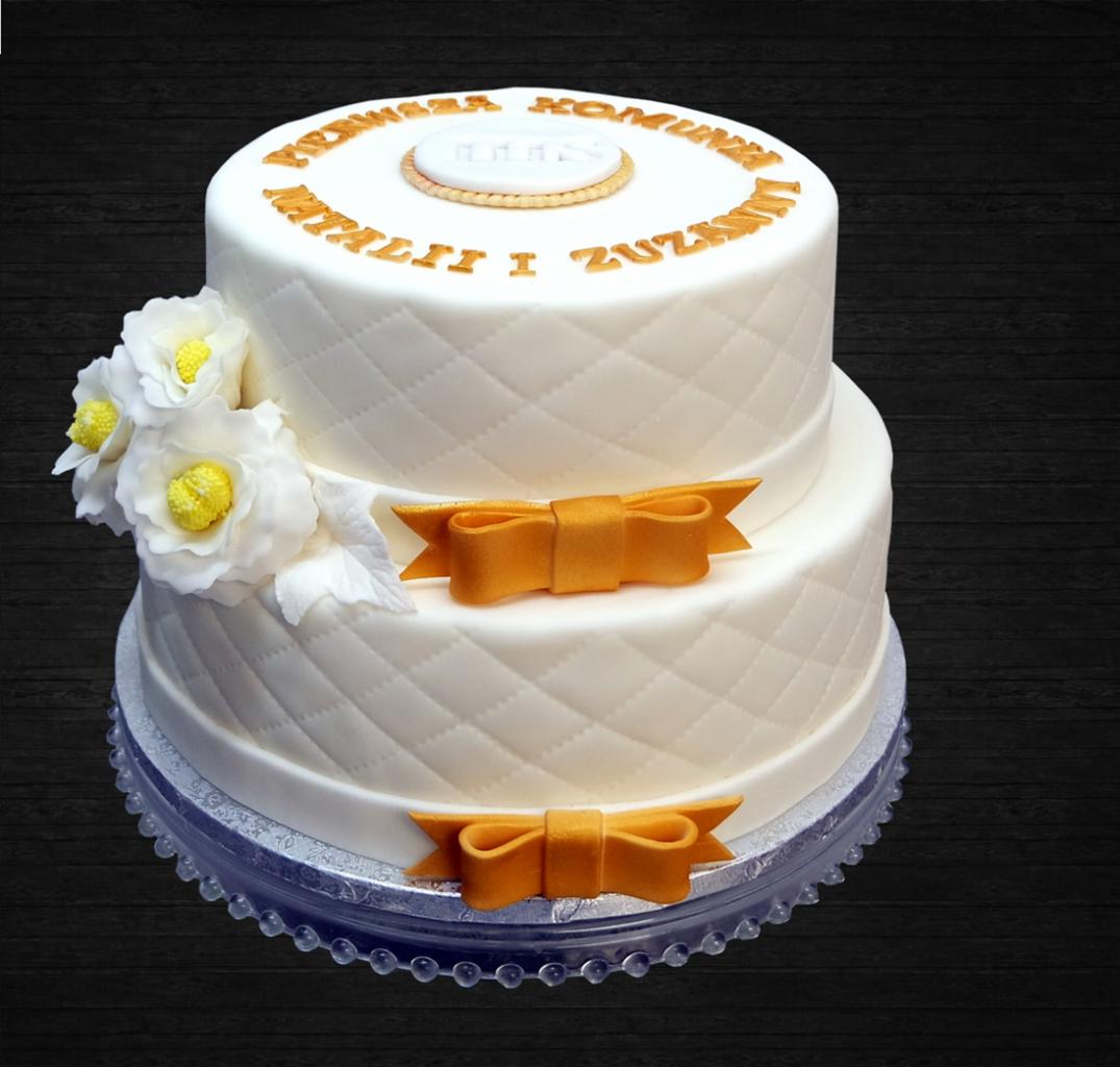 Dwupoziomowy tort komunijny z białym lukrem imitującym pikowanie, złotymi napisami 'Pierwsza Komunia Natalii i Zuzanny', dekoracjami z białych kwiatów i złotych kokardek, umieszczony...