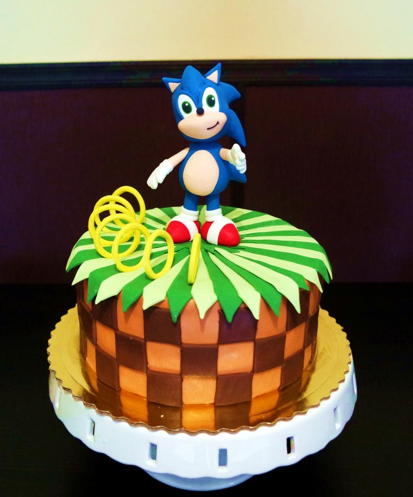Tort urodzinowy z motywem Sonic the Hedgehog, z figurką Sonica i żółtymi pierścieniami na zielono-brązowym tle w szachownicę, umieszczony na białym stojaku.