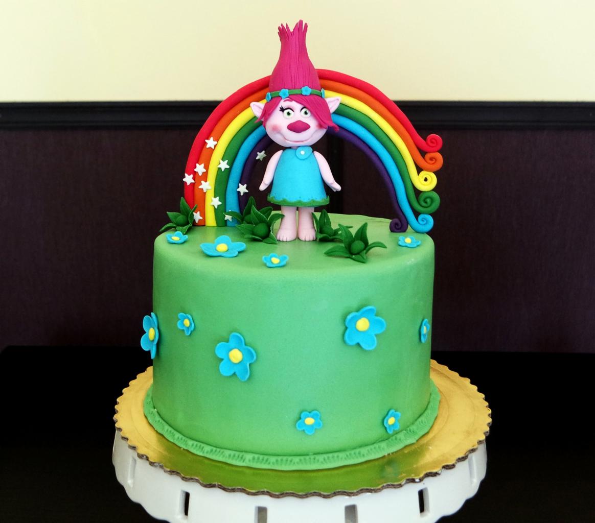 Zielony tort urodzinowy z figurką trolla Poppy pod tęczą, udekorowany niebieskimi kwiatkami i zielonymi listkami.