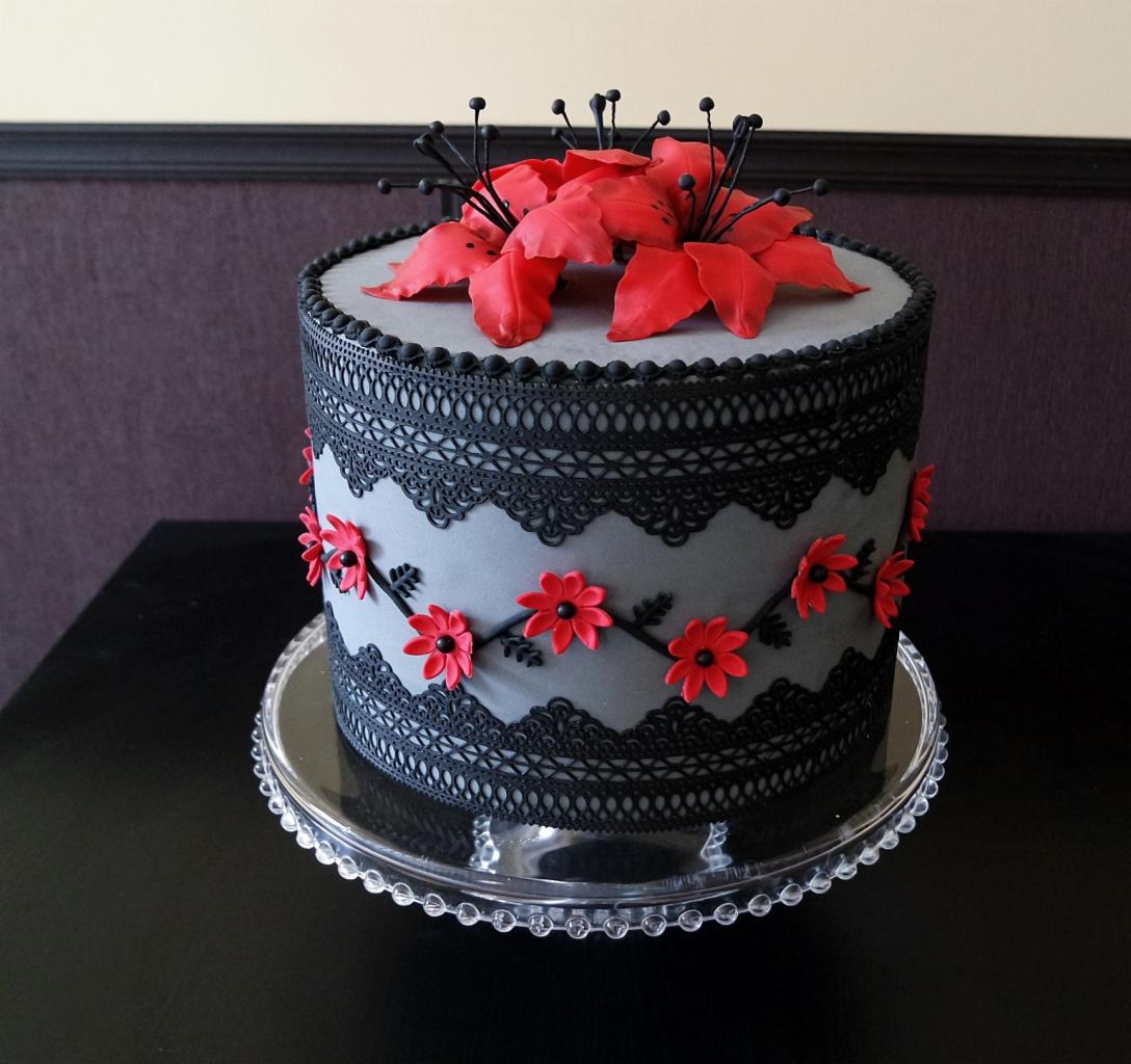Tort zdobiony czarną koronką i czerwonymi kwiatami na szarym tle, umieszczony na szklanej paterze.