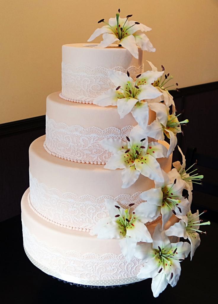 Czteropiętrowy tort w odcieniach pudrowego różu, dekorowany białymi koronkami i kaskadą lilii z zielonymi pręcikami.