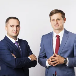 Portret dwóch uśmiechniętych mężczyzn w garniturach na jasnym tle, jeden z założonymi rękami, drugi z zegarkiem na nadgarstku.