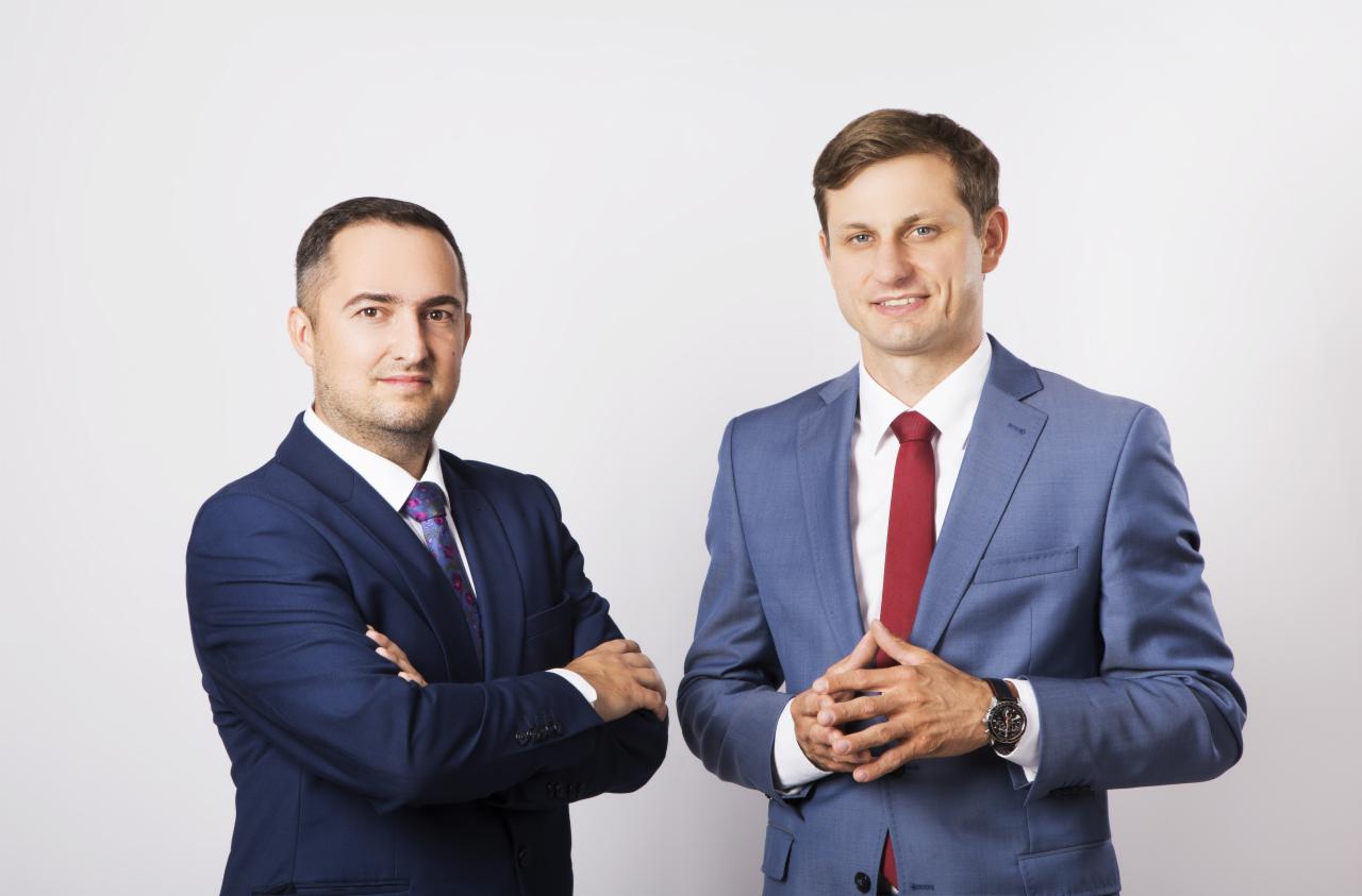 Portret dwóch uśmiechniętych mężczyzn w garniturach na jasnym tle, jeden z założonymi rękami, drugi z zegarkiem na nadgarstku.