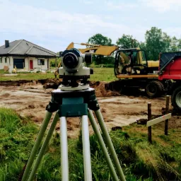 Geodezyjny niwelator optyczny na statywie na tle wykopu pod fundamenty domu, z koparką i wywrotką w tle.