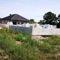 Budowa domu jednorodzinnego w trakcie wznoszenia ścian z białych bloczków Solbet, widoczne worki z materiałem budowlanym na murze, w tle drugi dom i drzewa.