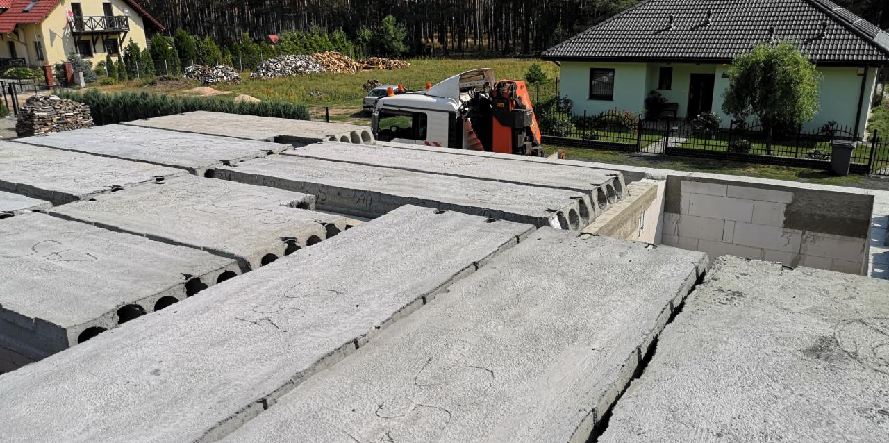 Montaż prefabrykowanych betonowych płyt stropowych z otworami, widoczny dźwig samochodowy i ściany z bloczków betonowych w tle.