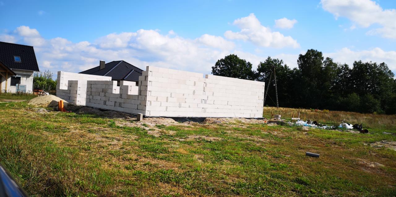 Wznoszone ściany z białych bloczków na fundamencie w trakcie budowy domu jednorodzinnego, widoczne elementy instalacji i sąsiednie budynki.